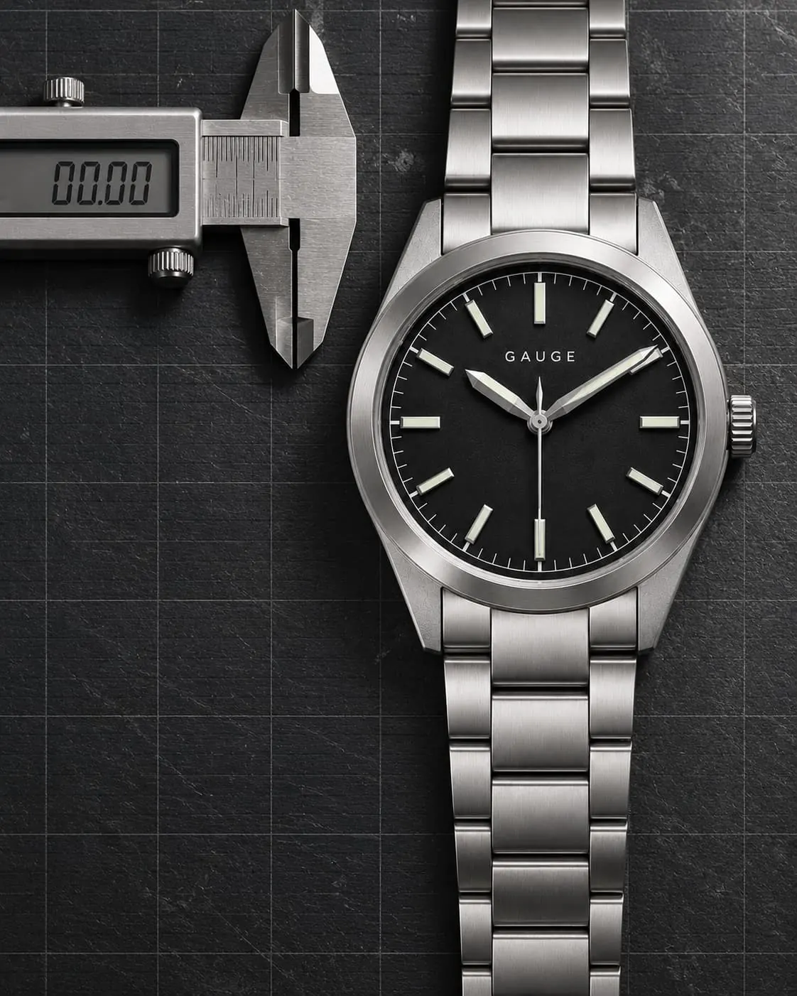 GAUGE-01 Field, black dial, steel bracelet, on a dark steel bench under a calibration grid beside a digital caliper reading zero.